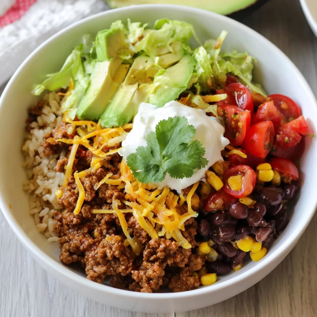 Close-up of a colorful Beef Burrito Bowl with brown rice, seasoned ground beef, avocado, corn, black beans, tomatoes, shredded cheese, and a dollop of sour cream.
