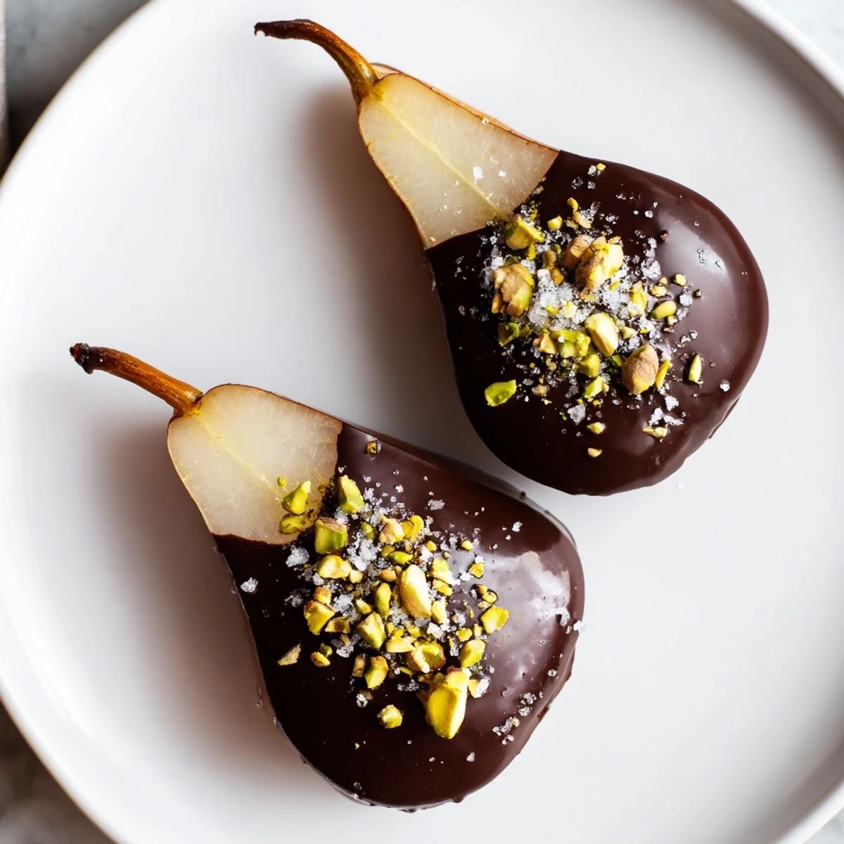 Juicy pear halves dipped in rich dark chocolate, sprinkled with sea salt and chopped pistachios on a parchment-lined tray.