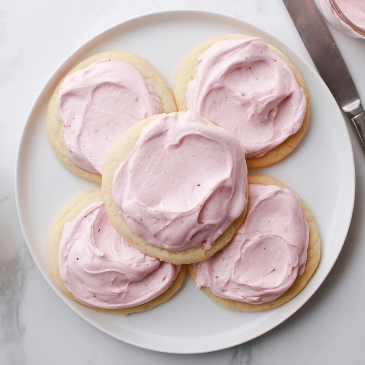 Golden Crumbl Sugar Cookies with thick, soft centers topped with vibrant pink almond frosting on a marble counter.